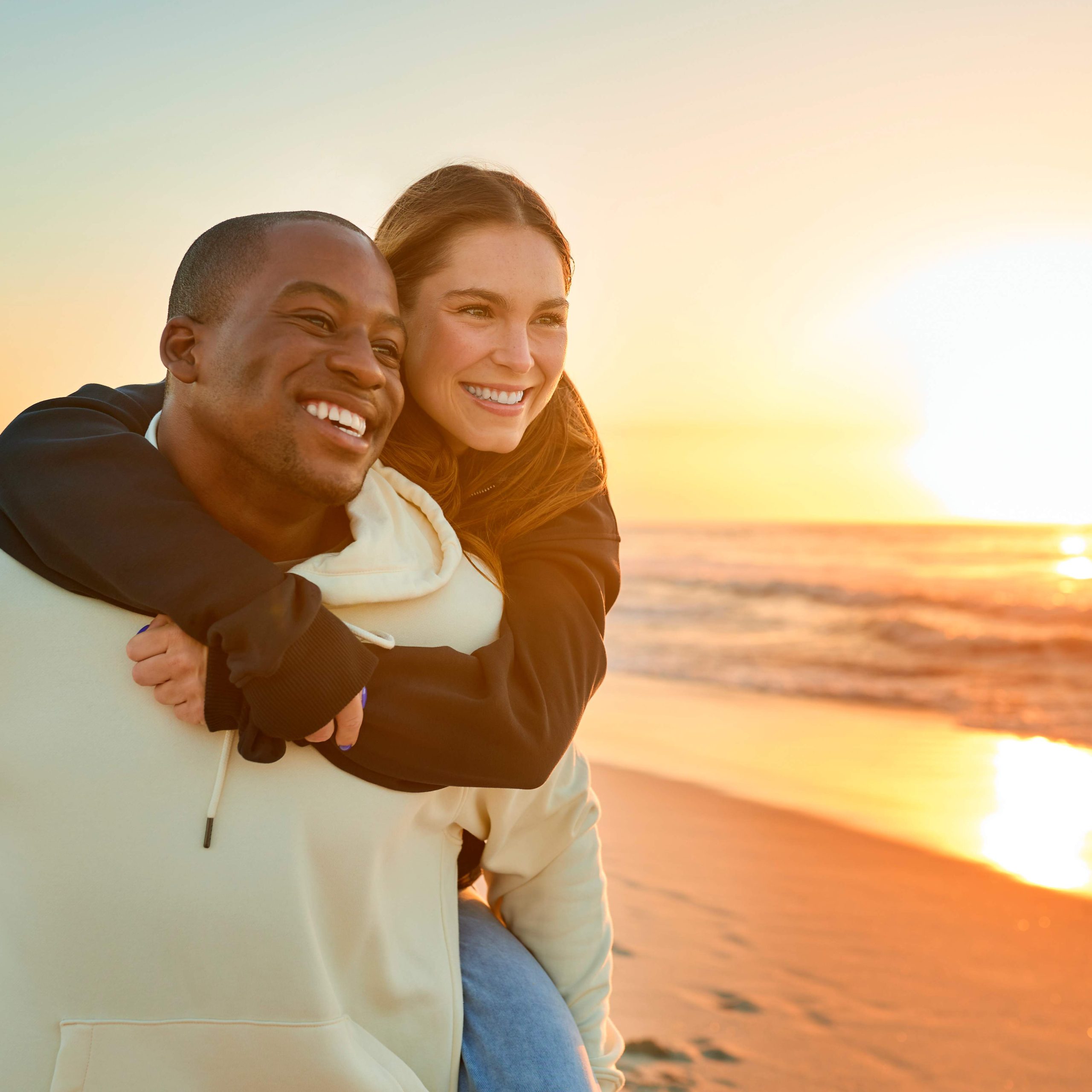 Casually Dressed Loving Couple With Man Giving Woman Piggyback Ride On Beach Shoreline At Sunrise