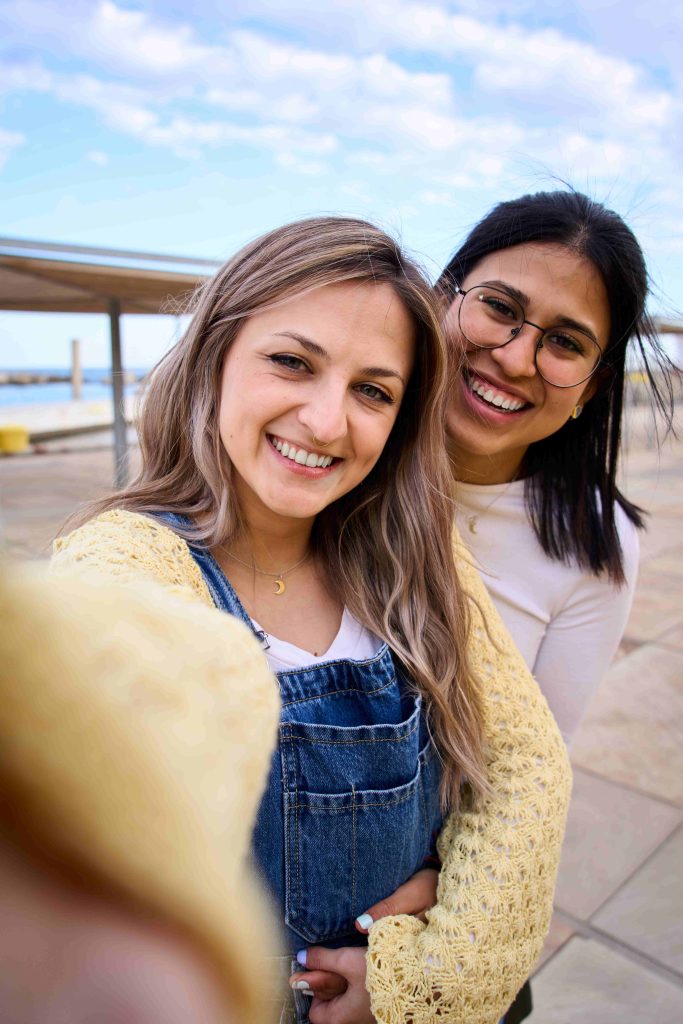 Vertical happy LGBT lesbian Caucasian couple on tourist vacation taking selfie phone promenade.